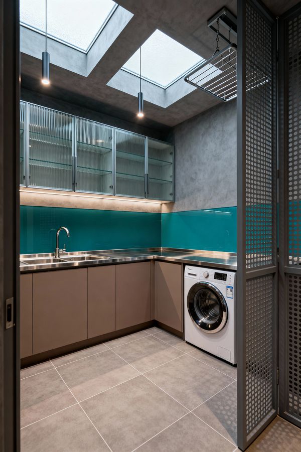 Compact modern laundry room with an L-shaped stainless-steel countertop and integrated sink, matte taupe handleless base cabinets, and a white front-loading washer. A glossy teal glass backsplash contrasts with gray microcement walls and large porcelain floor tiles, while fluted-glass upper cabinets, LED strip lighting, two skylights with slim pendants, a ceiling-mounted drying rack, and a perforated metal sliding screen complete the space.