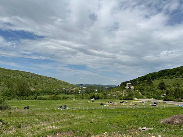 A scenic view of a green landscape with hills, vehicles parked in an open area, and a cloudy sky.