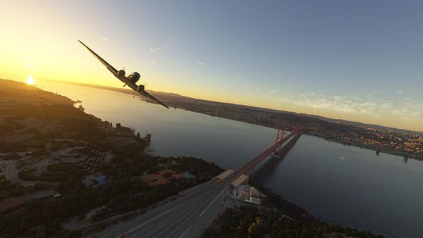 A vintage airplane flies over a river at sunset, with a prominent bridge in the background.