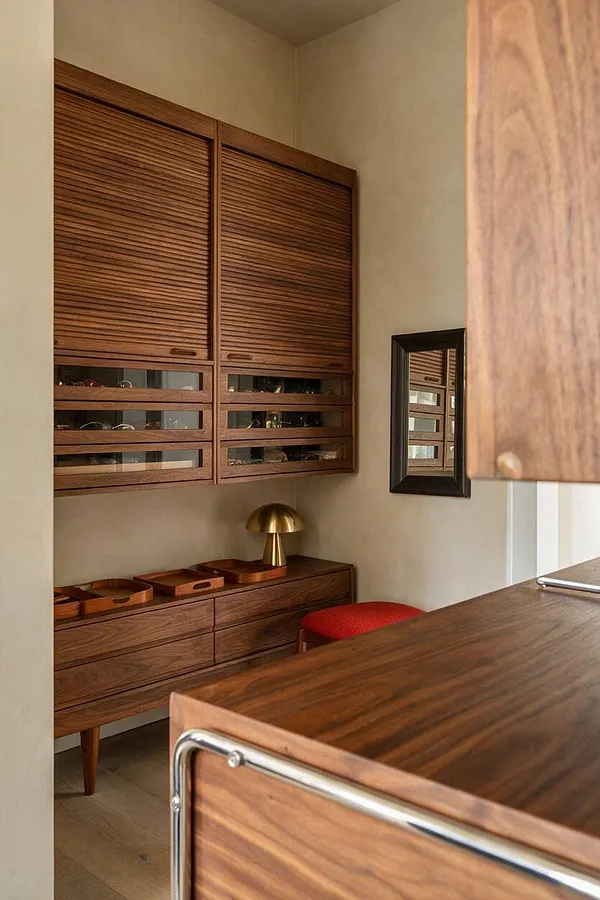 A compact mid-century modern closet features walnut slatted cabinet doors, glass-front storage, and a matching wood dresser beneath. A small brass mushroom lamp, a red upholstered stool, and a black-framed mirror add warmth and retro character against soft beige walls.