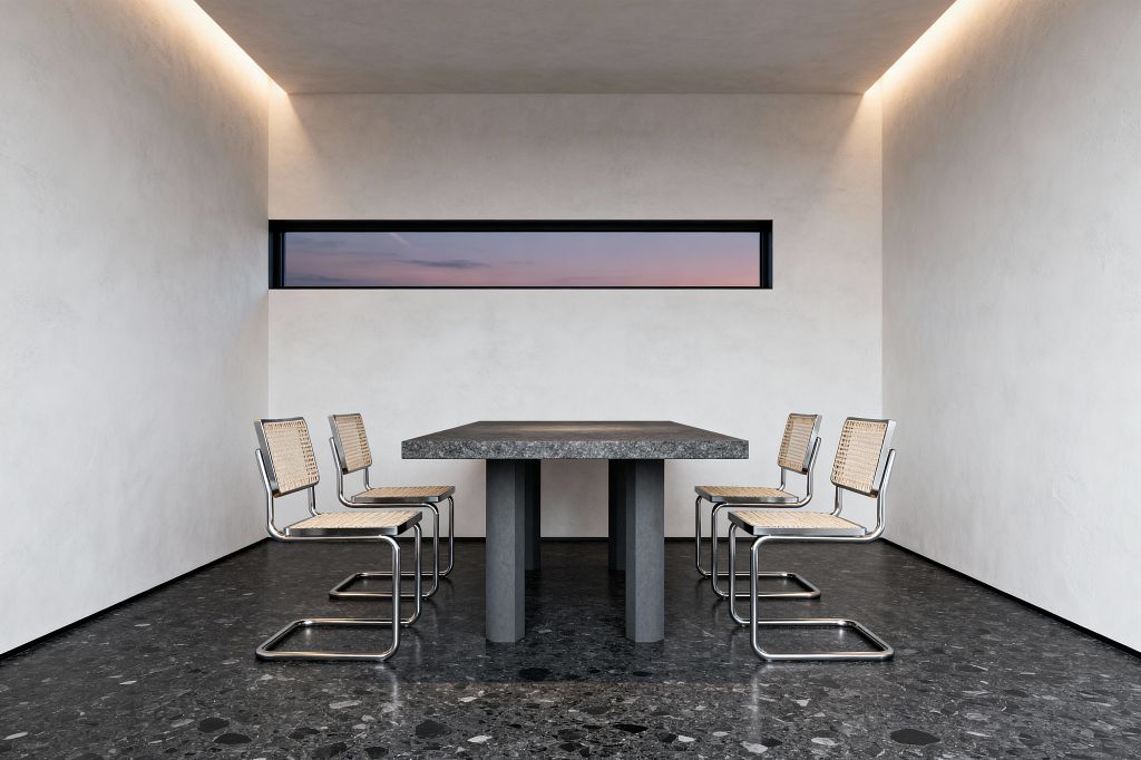 A minimalist dining room with a thick gray stone table on pedestal legs, surrounded by six chrome cantilever chairs with woven cane seats and backs. Smooth white plaster walls frame a narrow horizontal window showing a dusky sky, while warm cove lighting washes the ceiling. A dark charcoal terrazzo floor grounds the symmetrical, uncluttered arrangement, emphasizing clean lines and simple, tactile materials.