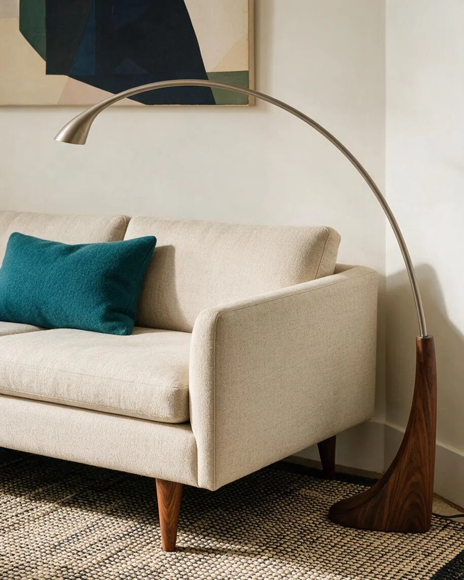 A Mid-Century Modern living room vignette with a cream upholstered sofa, a teal accent pillow, and a sweeping arc floor lamp with a wood base. The scene includes a textured woven rug, warm wood legs, and a minimalist abstract wall art piece above the sofa.