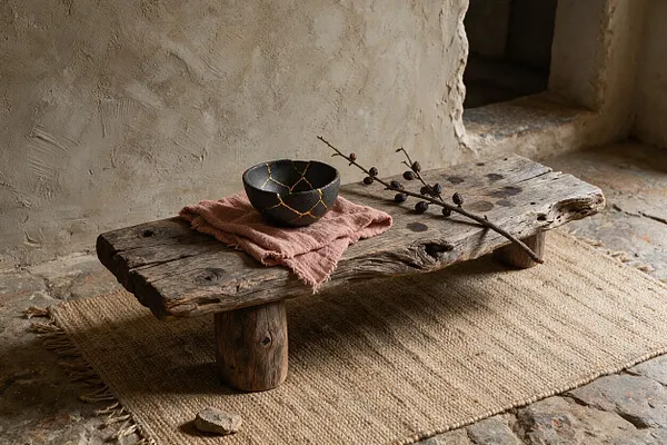 A low rustic wooden bench sits on a woven jute rug against a textured plaster wall. On top, a black bowl with gold kintsugi-style cracks rests on a dusty pink cloth beside a thin branch with seed pods, creating a calm, natural Wabi-Sabi still life.