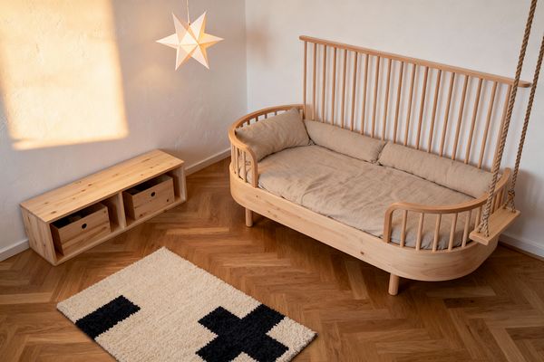A Scandinavian kids’ room with a light pine spindle-back daybed topped with beige linen cushions sits against a warm white textured wall. A white paper star pendant casts a soft glow above a low pine storage bench with two box drawers, while a natural-rope indoor swing hangs at the bed’s end; the space is grounded by honey-toned herringbone oak flooring and a cream high-pile rug with bold black cross motifs, all illuminated by late-afternoon sunlight.