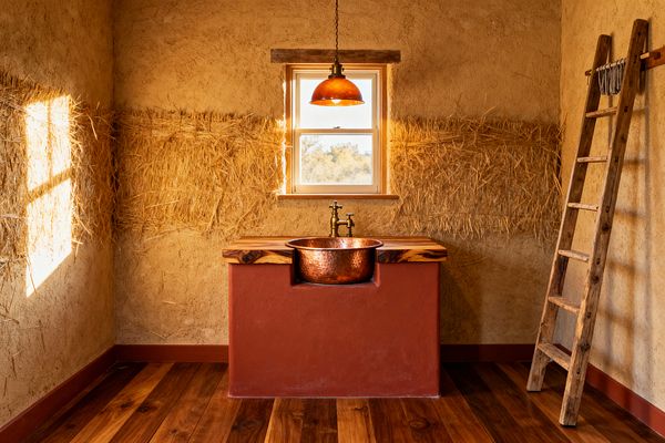 A small rustic laundry room features a centered window with a warm copper pendant above a hammered copper basin set into a terracotta base. A thick live-edge wood countertop and brass faucet sit against earthen plaster walls with a horizontal band of exposed straw. Wide-plank hardwood floors ground the space, and a weathered wooden ladder on the right serves as a drying rack. Sunlight casts golden tones and soft shadows across the walls and floor.