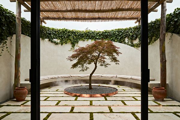 An enclosed patio with curved sand-colored stucco walls and a rustic pergola of timber posts and reed slats. At the center, a small Japanese maple rises from a copper-edged circle of black pebbles with soft mist. The floor is a grid of pale limestone pavers with green moss joints and a subtle linear light. A built-in curved bench with light beige cushions lines the wall, flanked by terracotta pots and trailing ivy.