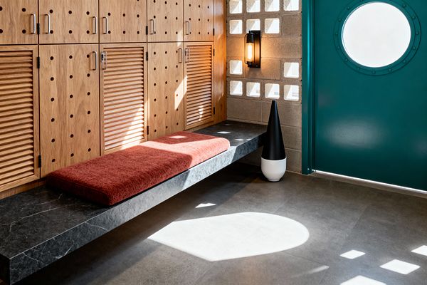 Mid-century mudroom with a wall of oak lockers featuring circular perforations and louvered doors, each with brushed metal pulls. A floating dark stone bench holds a rust-red cushion below a breeze-block wall and black sconce; opposite, a teal door with a round porthole window admits light onto large gray floor tiles, with a black-and-white cone-shaped decor piece nearby.