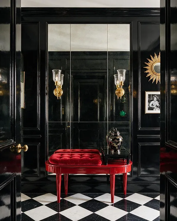 A dramatic Hollywood Regency hallway with glossy black paneled walls, a black-and-white checkered floor, and a bright red lacquer bench topped with a tufted velvet cushion. Gold sunburst accents, brass wall sconces, a mirrored back wall, and a small black side table with a decorative urn add glamour and contrast.