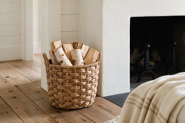 A cozy farmhouse interior detail showing a woven basket filled with chopped firewood beside a white plaster fireplace. Wide-plank wood flooring and a soft striped throw in the foreground add warmth and texture.