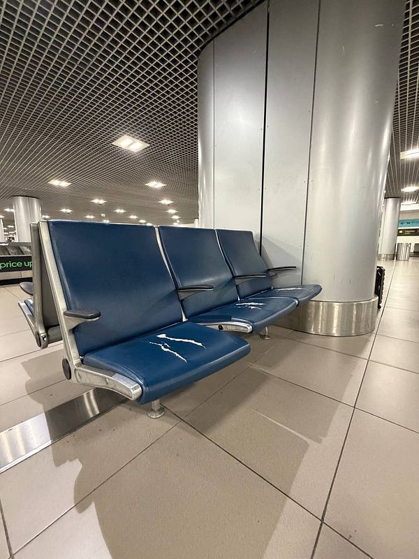 A row of three blue airport seats with worn and cracked surfaces in front of a large cylindrical pillar inside Lisbon Airport.