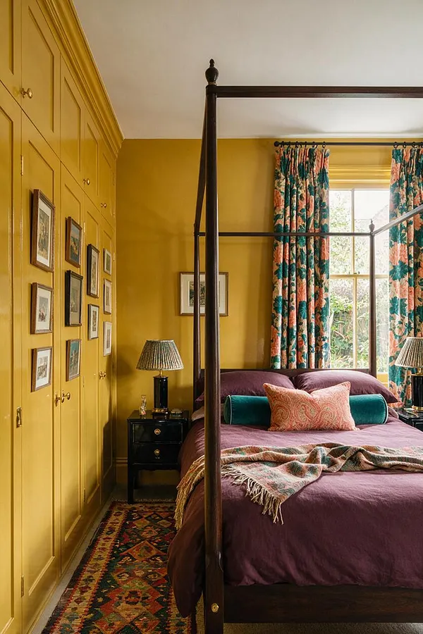 A maximalist master bedroom with mustard yellow walls and built-in wardrobes on the left, a dark wood four-poster bed centered in the room, and a plum duvet layered with teal and patterned accent pillows. Floral curtains frame a bright window, while small black bedside tables, table lamps, framed artwork, and a colorful patterned rug complete the richly layered look.
