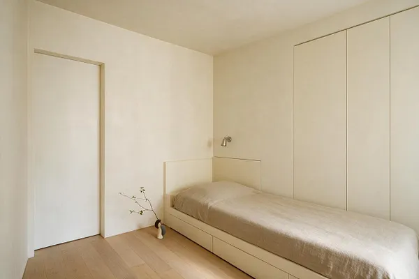 A minimalist guest bedroom with a single low bed dressed in beige bedding, built-in paneled storage along the wall, and a small wall-mounted reading light above the headboard. The room has white walls, light wood flooring, a plain door, and a simple vase with a bare branch adding a subtle decorative touch.