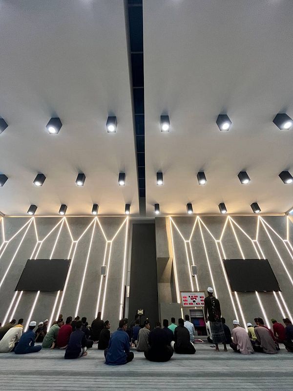 A group of people is seated in a modern mosque interior, preparing for prayer.