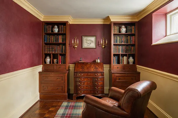 A traditional basement room styled as a cozy library and reading nook, with burgundy walls, cream wainscoting, crown molding, and a white ceiling. Two tall built-in bookcases flank a polished wood secretary desk, while a brown leather armchair sits on a hardwood floor with a plaid rug.