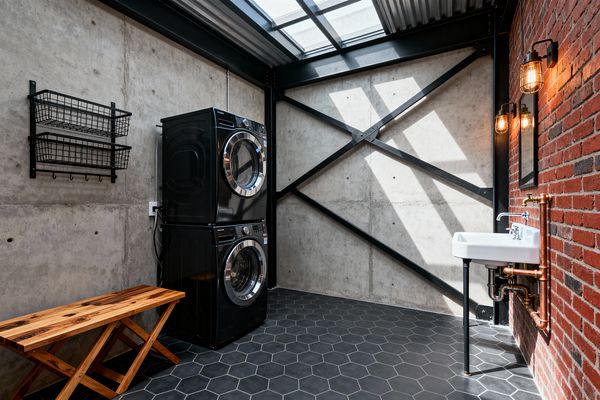 Industrial-style laundry room with raw concrete walls, a black steel X-brace, and a red brick wall; on the left, stacked black front-load washer and dryer sit beside two wire baskets and a simple wood bench. On the right, a white wall-mounted utility sink with exposed copper pipes, a small mirror, and caged sconces faces a charcoal hex tile floor, while a corrugated skylight above casts strong diagonal light and shadows.