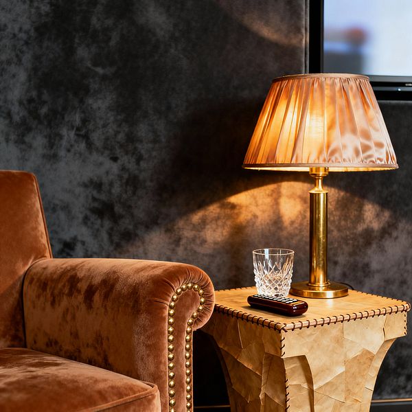 Close-up of a media room corner with a cognac velvet armchair featuring a rolled arm and brass nailhead trim. A stitched leather pedestal side table holds a cut crystal tumbler, a remote, and a brass table lamp with a pleated champagne shade. The lamp’s warm glow washes over dark charcoal, velvet-like walls, creating a moody, intimate atmosphere ideal for relaxed viewing.