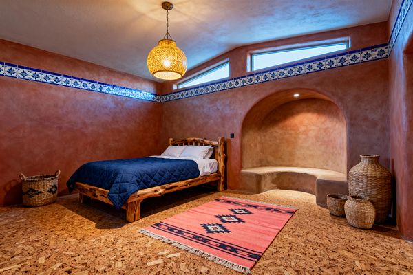 A Southwestern-inspired kids’ bedroom with terracotta plaster walls and a blue-and-white Talavera tile band near the ceiling. A rustic log bed with white sheets and a navy quilt sits on cork flooring next to a coral kilim rug; a curved adobe alcove with a built-in bench is lit by a recessed light, while a woven rattan pendant and natural baskets add warmth and texture beneath clerestory windows.