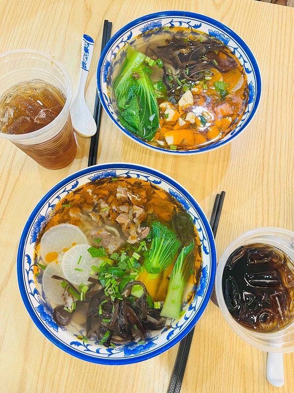 Two bowls of noodles with various toppings and drinks are presented on a wooden table.