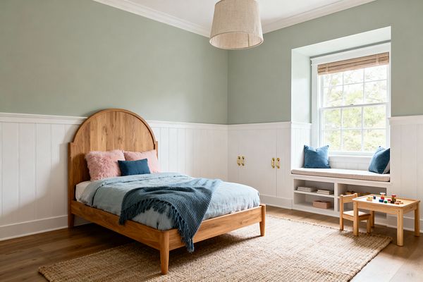 Bright kids’ bedroom with soft sage upper walls and white vertical wainscoting; a curved oak bed with light blue duvet, navy throw, and blush pillows sits on a large jute rug over warm wood floors. To the right, a deep window with a woven wood Roman shade features a cushioned bench with blue pillows and open cubbies below, brass pulls on nearby built-in cabinets, a simple linen drum pendant above, and a small oak play table with colorful blocks.