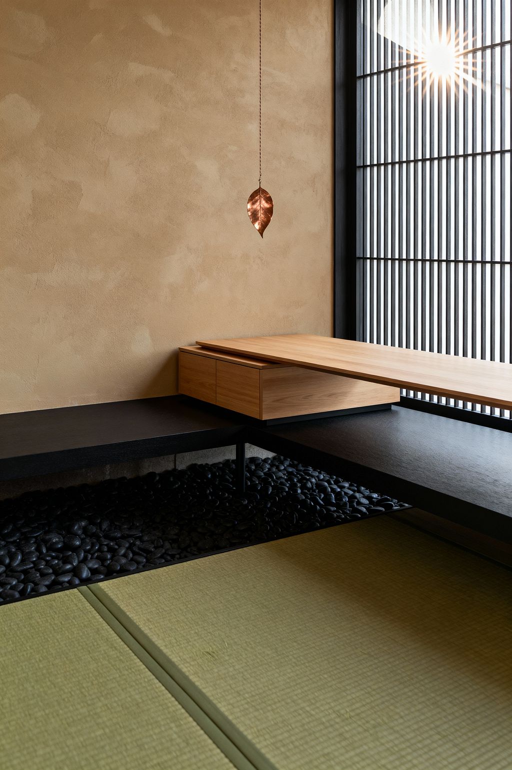 A minimalist Japanese-inspired home office with tatami mats, an ebonized L-shaped bench hovering over a bed of black river stones, and a floating light oak desk with concealed drawers. A warm sand-colored clay-plaster wall and a black slatted shoji-style window filter sunlight, while a small copper leaf pendant hangs on a chain, creating a calm, uncluttered workspace for floor-level seating and focused tasks.