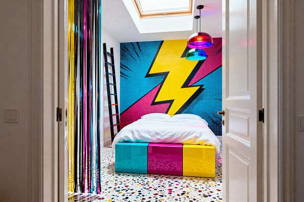 A kids’ bedroom viewed through white double doors showcases a bold pop art mural with a large yellow lightning bolt over blue halftone dots and magenta shapes. A bed with a glossy cyan, magenta, and yellow base sits on a multicolor terrazzo floor beneath a skylight and two multicolored glass pendant lights; a black leaning ladder and metallic CMYK foil streamers add playful, graphic accents.