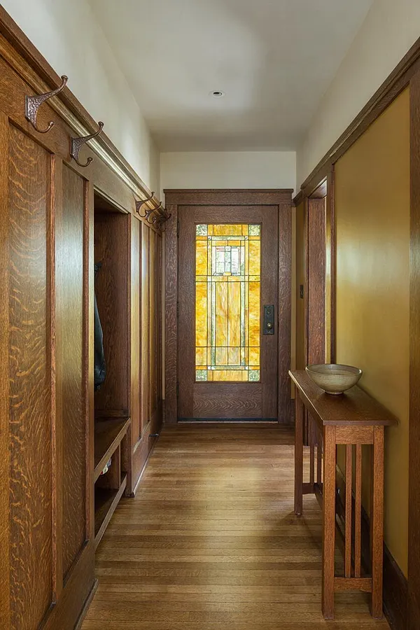 A narrow Craftsman-style entryway with rich wood trim, built-in coat storage on the left, and a slim console table with a bowl on the right. At the end is a wooden front door with a tall stained-glass panel in amber and green tones, creating a warm, traditional focal point.