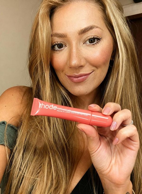 A woman is smiling while holding a tube of lip product in a bathroom setting.