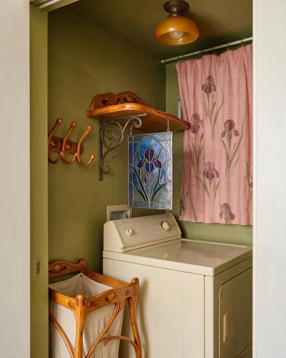 A compact laundry room features a cream top-loading washer against olive green walls, with a pink floral curtain, stained glass iris panel, and carved wooden shelves and hamper. Curving Art Nouveau details in the wall hooks, woodwork, and lamp create a decorative, vintage-inspired look.