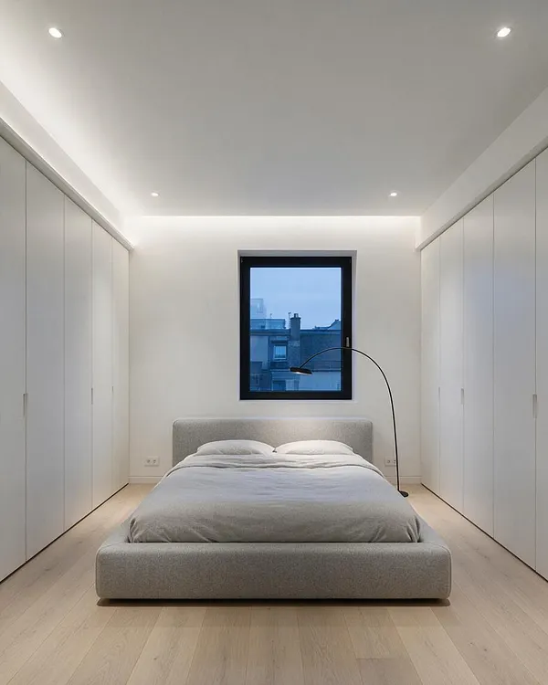 A serene minimalist master bedroom with a low upholstered platform bed centered under a black-framed window, flanked by full-height white built-in wardrobes on both sides. The room features light wood flooring, crisp white walls, recessed ceiling lights, and a slender arched floor lamp for a clean, modern look.