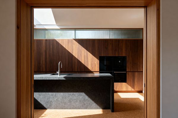 View through a wood-framed opening to a minimalist kitchen with a dark charcoal stone island featuring an undermount sink and stainless gooseneck faucet. A full wall of flat-panel walnut cabinets spans the back, interrupted by stacked black built-in ovens. Above, a clerestory strip of fluted glass and a skylight pour diagonal sunlight across the space. The floor is warm cork, and the palette is restrained in walnut brown, charcoal gray, black, and soft white.
