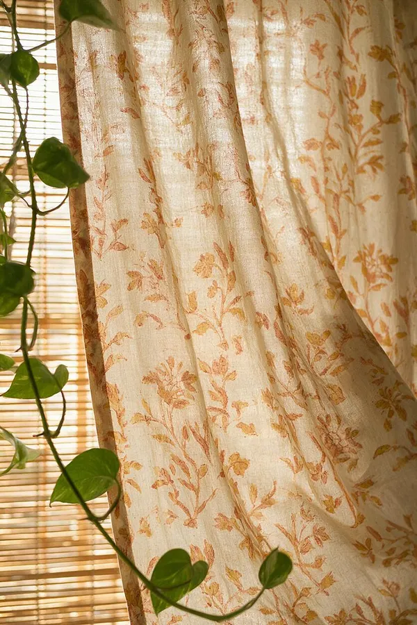 A close-up of sheer cream curtains with a soft orange floral vine print, glowing in warm sunlight. A trailing green houseplant hangs in the foreground, and woven bamboo blinds filter the light behind the fabric.