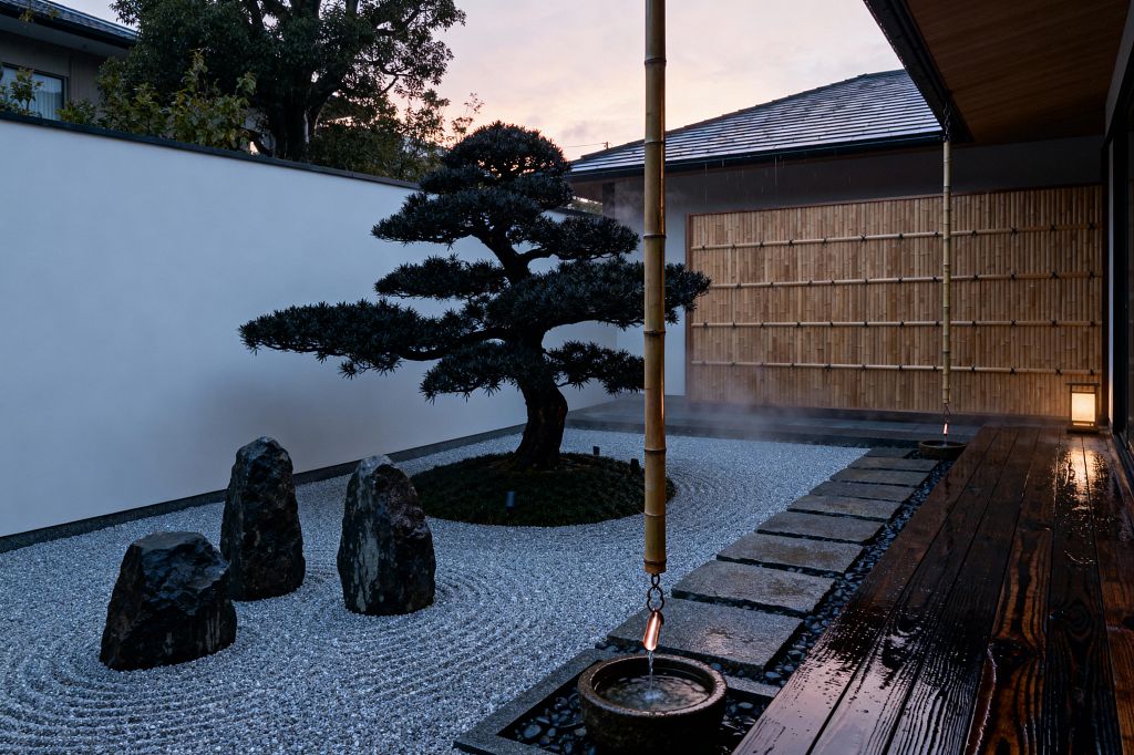 A serene Japanese Zen patio centers on a carefully pruned pine on a moss mound, encircled by raked white gravel and three dark standing stones. A bamboo rain chain drips into a small stone basin beside a wet, dark-stained wood deck, with granite stepping stones, a smooth white plaster wall, and a honey-colored bamboo screen; a soft lantern glows as evening mist gathers, creating a calm, contemplative mood.