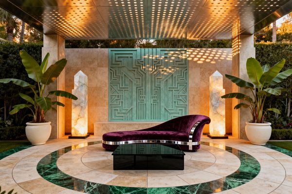 A symmetrical outdoor seating area with a plum velvet chaise lounge and low black glass coffee table sits on a circular floor of travertine and emerald marble. Behind, a mint green Art Deco relief panel is set into beige stone, flanked by two tall glowing alabaster obelisk lights and large banana plants in cream urns. An overhead perforated metal canopy casts dotted sunlight across the scene.