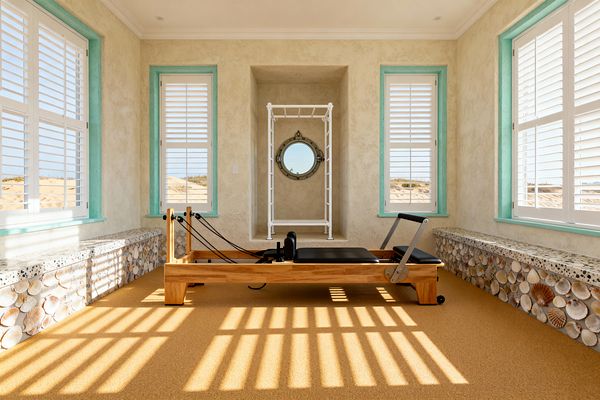 Bright home gym with a light-wood Pilates reformer centered between windows fitted with white plantation shutters and aqua trim. Sandy textured stucco walls and a golden cork floor set a beachy palette. Built-in side benches have terrazzo tops and fronts covered in assorted seashells. At the far wall, a white stall bar frames a metal-rim porthole mirror. Sunlight streams through the shutters, casting striped shadows across the room.