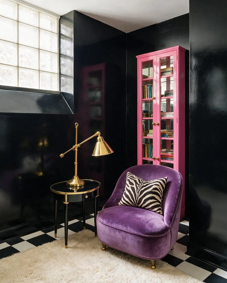 A dramatic basement sitting area with glossy black walls, a black-and-white checkered floor, and a frosted glass block window bringing in light. A plush purple velvet chair with a zebra-print pillow sits beside a black round side table with a brass lamp, while a bright pink cabinet with glass doors displays books in the corner.