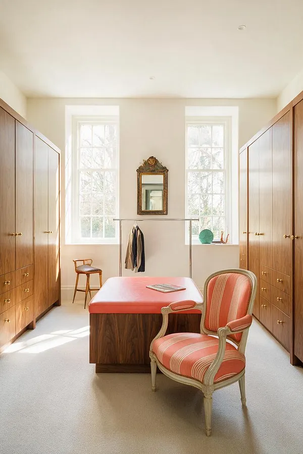 A bright eclectic closet with tall wood wardrobes lining both sides, a coral upholstered bench in the center, and a striped vintage armchair in the foreground. Two tall windows bring in natural light, and a decorative antique mirror hangs above a slim garment rack at the back wall.