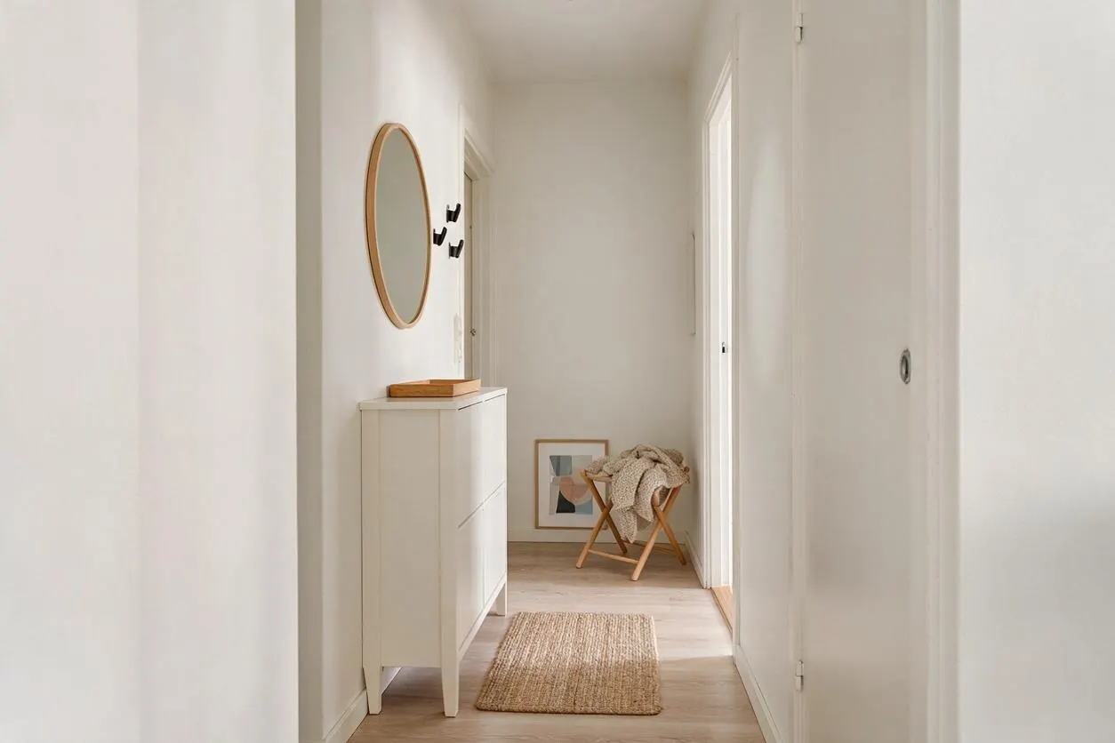 A narrow Scandinavian-style entryway with white walls, light wood flooring, and a soft neutral rug. A slim white cabinet with a round wood-framed mirror sits on the left, while a small wooden stool with a draped knit throw and a framed print add warmth at the end of the hall.