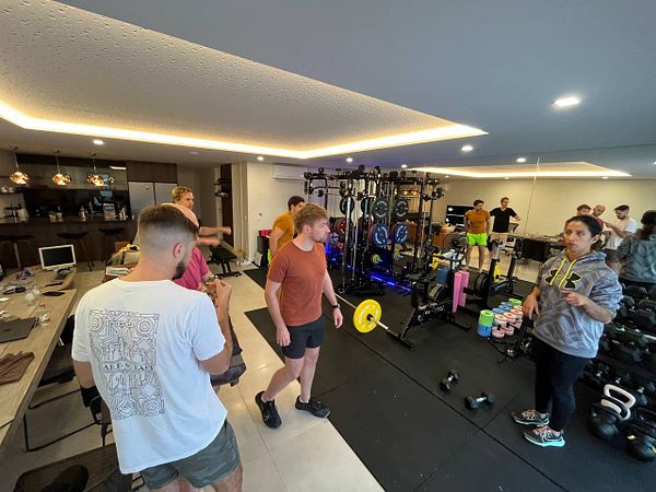 A group workout session is taking place in a home gym setting with several participants engaged in conversation and exercise.