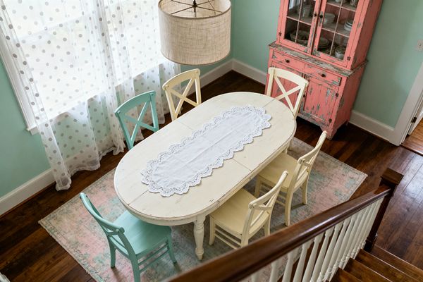 A bright dining room with mint green walls centers on an oval, distressed antique-white table topped with a scalloped lace runner and surrounded by mismatched cross-back chairs in buttercream and soft aqua on a faded pastel rug. A coral-pink weathered hutch with glass doors displays white dishes as sheer polka-dot curtains diffuse daylight, a beige linen drum pendant hangs above, and warm hardwood floors with white trim complete the cozy shabby chic look.