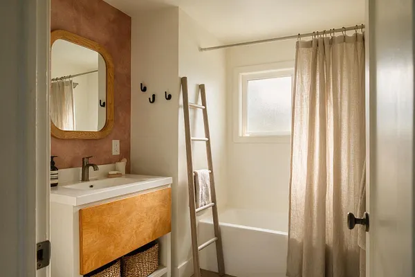 A small Organic Modern bathroom with a white vanity, warm wood cabinet front, rounded wood-framed mirror, and terracotta accent wall behind the sink. A light linen shower curtain, small frosted window, black hooks, and a wooden ladder add natural texture and a calm, spa-like feel.