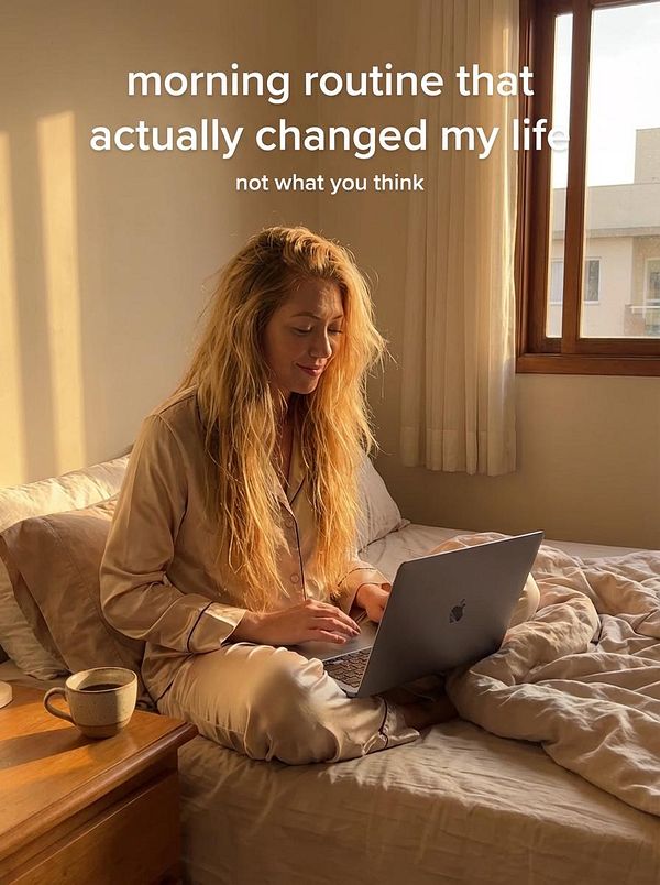 A woman in pajamas sits on a bed, working on a laptop in a sunlit room.