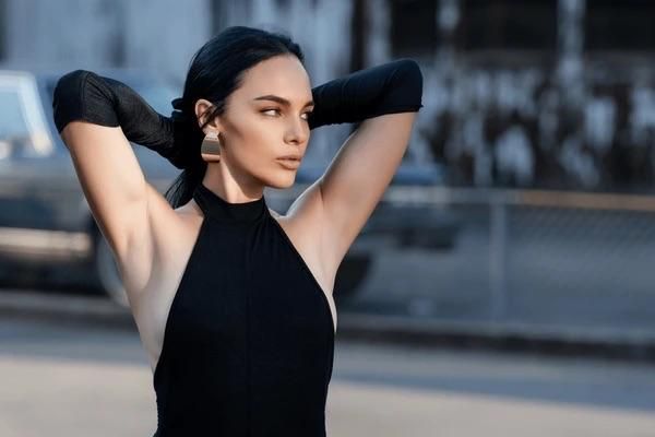 A woman poses confidently outdoors, showcasing a stylish black outfit and striking features.