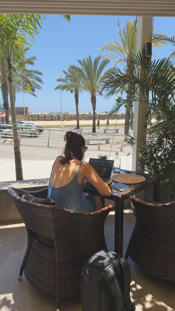 A person is working on a laptop at a beachside café under palm trees.