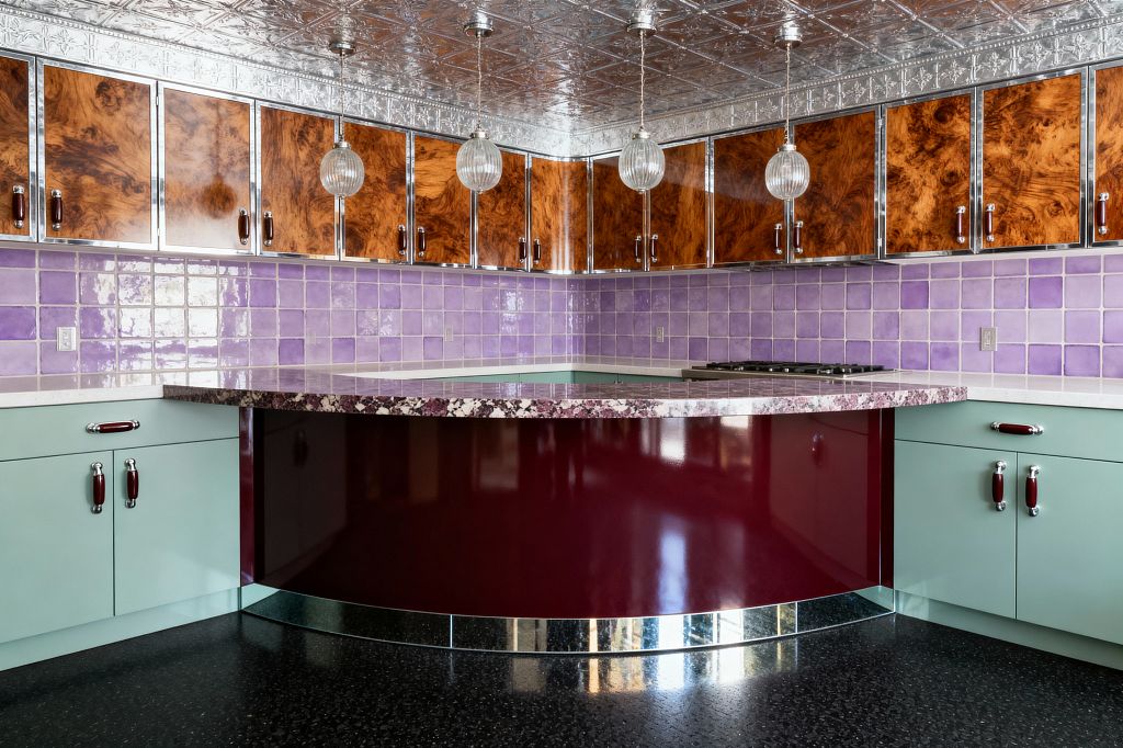 A vintage glam kitchen centers on a curved, high-gloss burgundy peninsula beneath three ribbed glass globe pendants. Lilac ceramic tile lines the backsplash, mint green base cabinets with chrome and maroon pulls sit below glossy burl wood uppers edged in chrome, a pressed-tin silver ceiling and mirrored toe-kick add sparkle, while a purple-flecked stone countertop, gas cooktop, and black speckled floor complete the retro-luxe space.