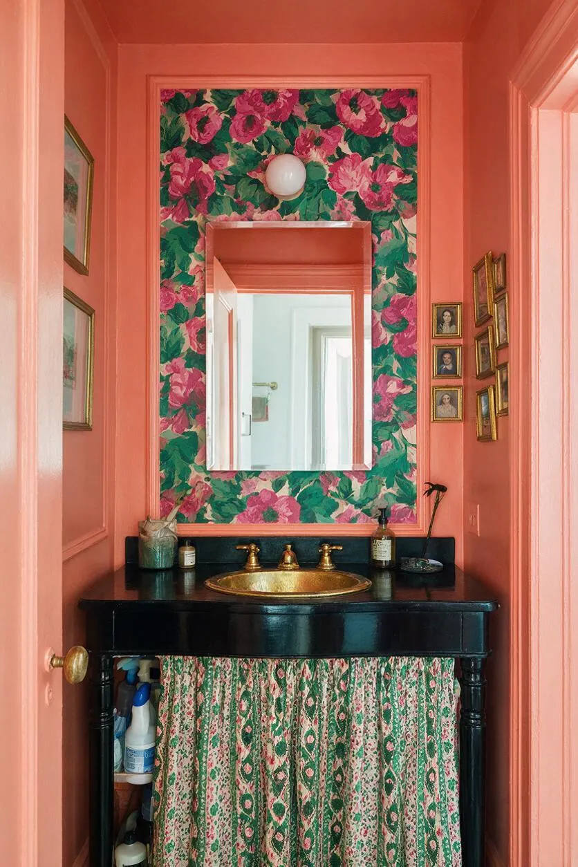 A narrow powder room painted in coral pink with a large floral wallpaper panel framing a rectangular mirror above a black vanity. The vanity has a gold faucet and sink, patterned fabric curtain below, and small framed portraits and decor clustered on the side walls.