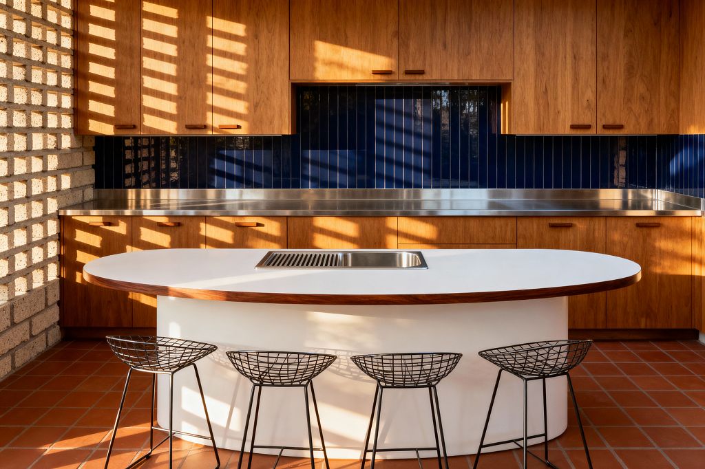 A mid-century modern kitchen features flat-front walnut cabinetry around a stainless worktop and a glossy navy backsplash of vertical tiles. In front, a white oval island with a walnut edge band includes a centered rectangular stainless insert and is paired with four black wire barstools; terracotta floor tiles and a pale brick screen wall catch sunlight, casting geometric shadows across the warm wood and deep blue surfaces.
