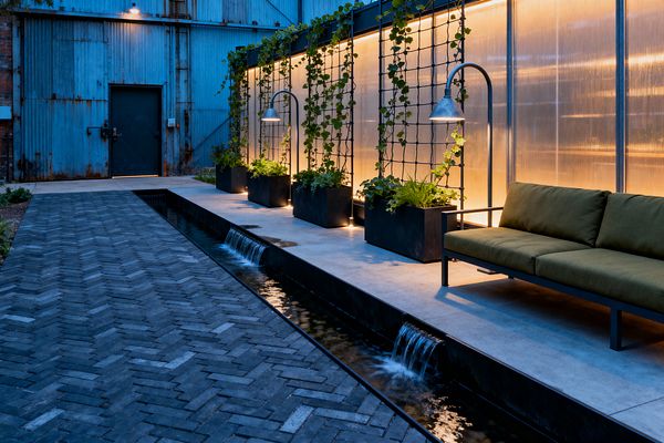 An industrial patio sits beside a weathered blue corrugated-metal building. A long concrete platform borders a narrow water rill with two small waterfalls, while charcoal herringbone brick paving runs alongside. Black steel planters with grid trellises support climbing vines in front of a glowing translucent polycarbonate wall. Curved galvanized lamps hover over the greenery, and a low sofa with olive cushions completes the warmly lit, urban setting.