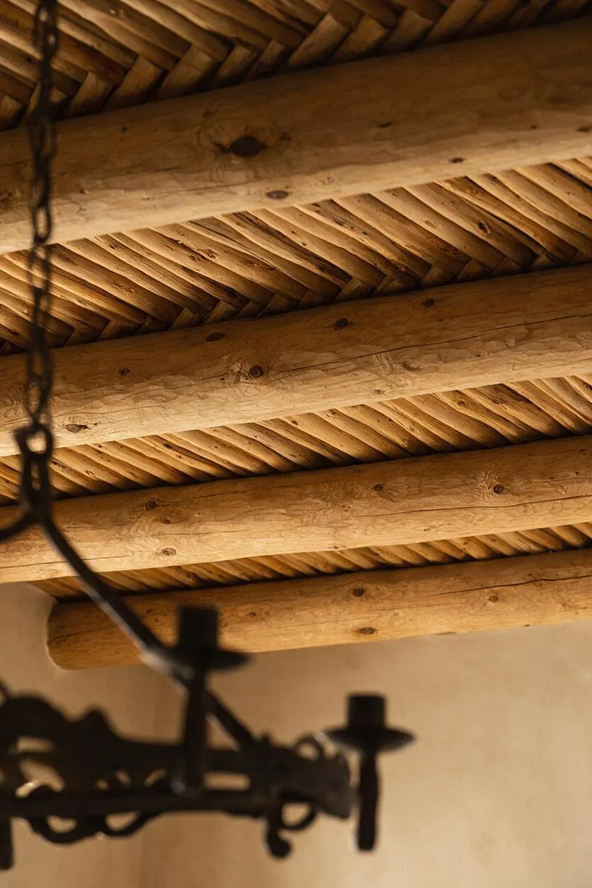 A close-up view of a Southwestern-style ceiling showing thick exposed log beams and a woven reed or branch infill pattern between them. A dark wrought-iron light fixture hangs out of focus in the foreground against a warm beige wall.