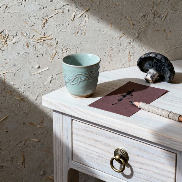 Close-up of a Wabi-Sabi home office vignette: a whitewashed wood desk with a small drawer and brass ring pull sits beside a rough clay-plaster wall embedded with straw. On the desktop are a pale celadon ceramic cup, a brown card with black ink strokes, a pencil wrapped in natural twine, and a dark mushroom, with angled sunlight casting soft shadows across the textured surfaces.