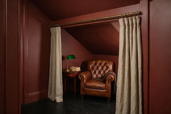 A compact basement reading nook framed by beige curtains, with deep burgundy walls and a sloped ceiling. Inside sits a tufted brown leather armchair beside a small wooden side table topped with a green banker’s lamp and a stack of books, creating a cozy Dark Academia atmosphere.